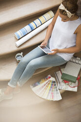 Frau mit digitalem Tablet auf einer Treppe, umgeben von Tapeten und Farbmustern - CAIF17212