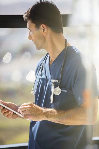 Männlicher Arzt mit Tablet-PC und Blick aus dem Krankenhausfenster, lizenzfreies Stockfoto