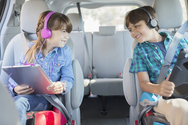 Fröhliche Geschwister mit Kopfhörern und digitalen Tablets auf dem Rücksitz eines Autos - CAIF17689