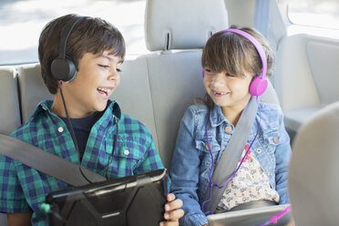 Happy brother and sister with headphones using digital tablets in back seat of car - CAIF17691