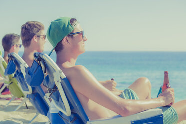 Junge Männer trinken Bier in Liegestühlen am sonnigen Strand - CAIF17699