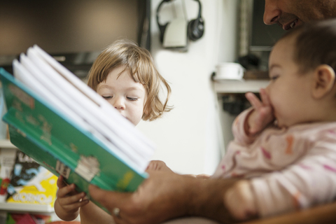Ausgeschnittenes Bild eines Vaters, der ein Buch hält, während er mit seinen Töchtern zu Hause sitzt, lizenzfreies Stockfoto