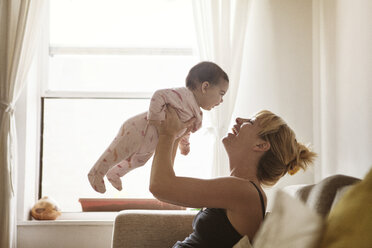Happy mother lifting baby girl while sitting on sofa at home - CAVF09172