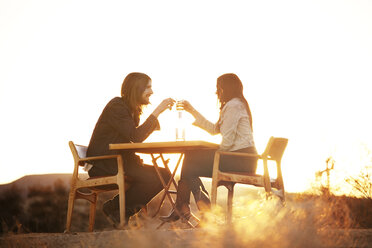 Paar stößt mit Weingläsern an, während sie auf Stühlen auf einem Feld sitzen - CAVF09337