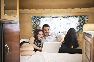 Lächelndes Paar, das einen Laptop benutzt, während es im Wohnmobil im Bett liegt - CAVF09406