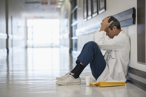 Arzt auf dem Boden sitzend in einem Krankenhauskorridor mit dem Kopf in den Händen, lizenzfreies Stockfoto