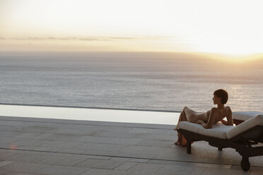 Frau im Kleid auf Liegestuhl auf der Terrasse mit Blick auf das Meer bei Sonnenuntergang - CAIF19060