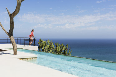 Frau steht auf dem Balkon am Pool mit Blick auf das Meer - CAIF19096