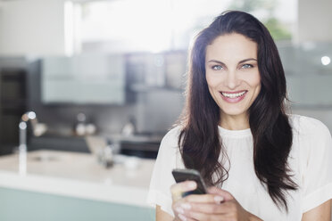 Porträt einer lächelnden Frau, die in der Küche eine SMS mit ihrem Mobiltelefon schreibt - CAIF19119