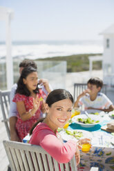 Familie beim Mittagessen am Tisch auf der sonnigen Terrasse mit Blick aufs Meer - CAIF19298