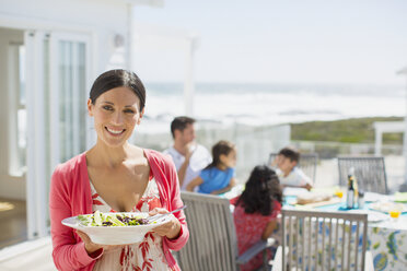 Frau hält Salatschüssel auf sonniger Terrasse mit Blick aufs Meer - CAIF19299