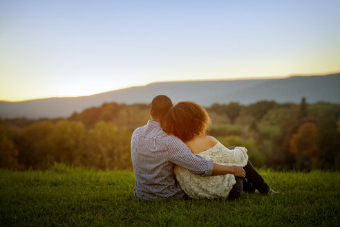 Rückansicht eines Paares, das bei Sonnenuntergang auf einer Wiese sitzt - CAVF09566