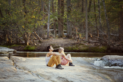 Paar schaut auf, während es auf einem Felsen am Fluss im Wald sitzt, lizenzfreies Stockfoto