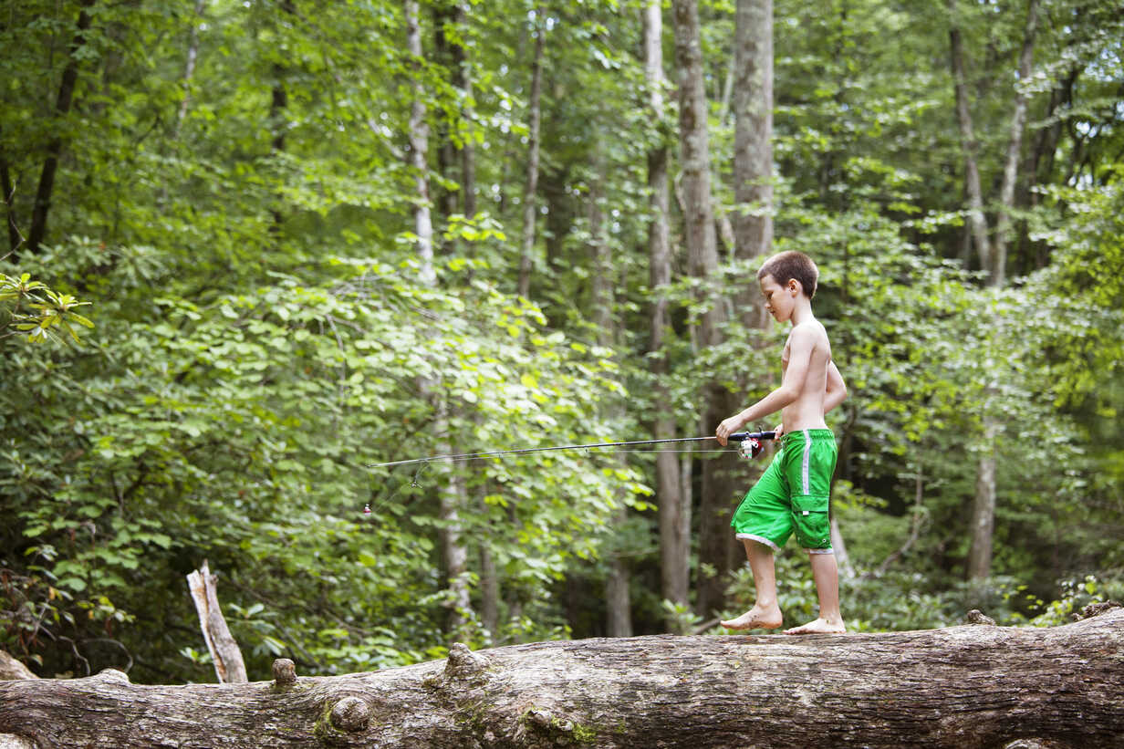 Side view of boy with fishing rod walking on fallen tree trunk in forest  stock photo