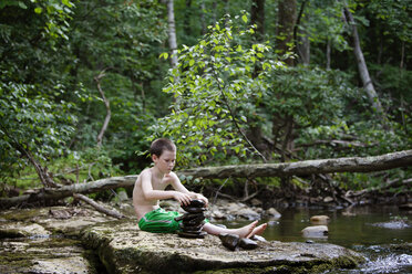 Junge arrangiert Kieselsteine, während er auf einem Felsen am Seeufer sitzt - CAVF09873