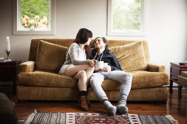 Couple holding coffee cups while sitting on sofa at home - CAVF09955