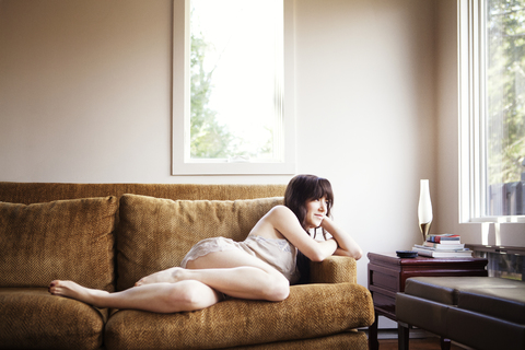 Nachdenkliche Frau, die wegschaut, während sie sich auf dem Sofa zu Hause entspannt, lizenzfreies Stockfoto