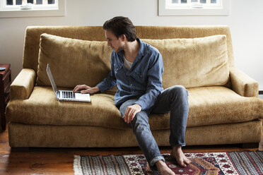 Man using laptop computer while sitting on sofa at home - CAVF09979