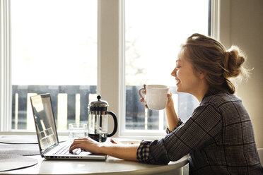 Glückliche Frau, die einen Laptop benutzt, während sie einen Kaffee am Tisch genießt - CAVF10037