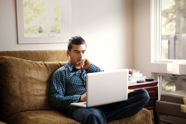 Mann benutzt einen Laptop, während er zu Hause auf dem Sofa sitzt - CAVF10038