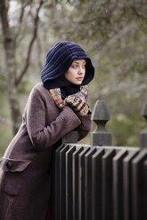 Frau in warmer Kleidung schaut weg, während sie sich an einen Zaun lehnt - CAVF10059