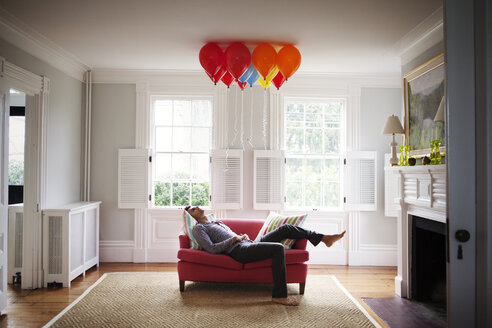 Man looking at helium balloons while lying on sofa - CAVF10121