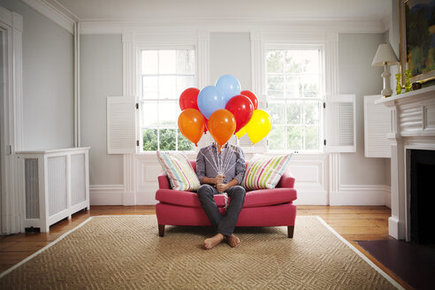 Man holding helium balloons while sitting on sofa at home - CAVF10122