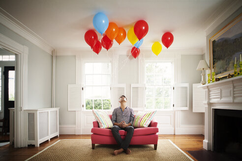 Man looking up at balloons while sitting on sofa at home - CAVF10123