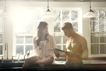 Mann liest Zeitung, während seine Freundin mit einer Kaffeetasse auf dem Küchentisch sitzt - CAVF10124