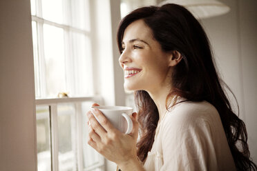 Lächelnde Frau mit Kaffeetasse, die durch ein Fenster zu Hause wegschaut - CAVF10128