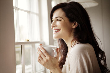 Smiling woman with coffee cup looking away through window at home - CAVF10128