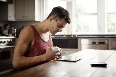 Mann mit Kaffeetasse schaut auf Tablet-Computer, während er am Tisch in der Küche sitzt - CAVF10131