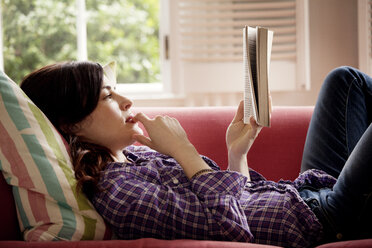 Nachdenkliche Frau liest ein Buch, während sie zu Hause auf dem Sofa liegt - CAVF10140