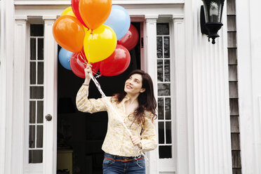 Woman holding helium balloons while standing outside house - CAVF10147