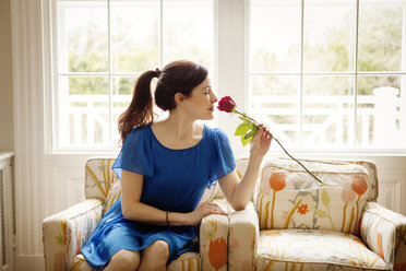 Frau riecht an einer roten Rose, während sie zu Hause auf einem Sessel sitzt - CAVF10150