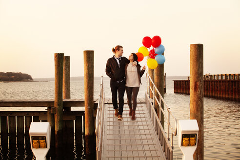 Frau hält Heliumballons, während sie mit einem Mann auf dem Pier bei Sonnenuntergang spazieren geht - CAVF10215