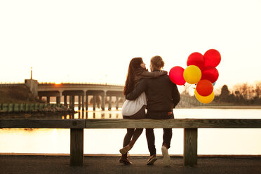 Rear view of couple with balloons sitting on railing during sunset - CAVF10216