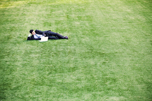 Businessman looking at laptop computer while lying on grassy field - CAVF10252