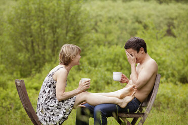 Couple holding coffee cups while sitting on chair in field - CAVF10268