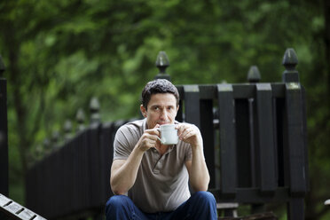 Man with coffee cup sitting by fence - CAVF10275