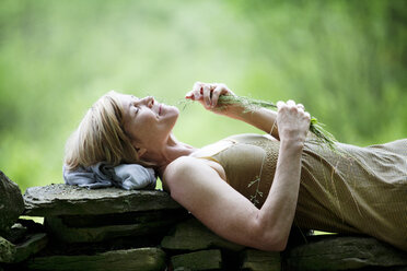 Woman playing with grass while lying on stone wall - CAVF10277