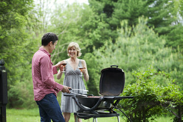 Smiling couple having drink while preparing food on barbecue - CAVF10290