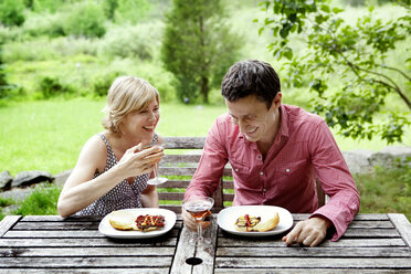 Happy couple having food while sitting by table on field - CAVF10295