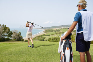 Caddy beobachtet Frau beim Abschlag auf dem Golfplatz mit Blick aufs Meer - CAIF19489