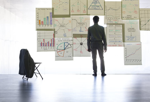 Geschäftsmann betrachtet Diagramme und Tabellen an einer Glaswand im Büro - CAIF19674