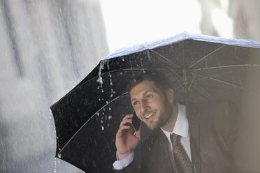 Geschäftsmann telefoniert unter Regenschirm im Regen - CAIF19707