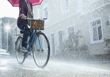 Frau fährt Fahrrad mit Regenschirm in regnerischer Straße - CAIF19712