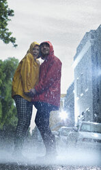 Glückliches Paar in Regenmänteln hält sich im Regen an den Händen - CAIF19732
