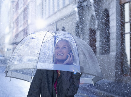 Lächelnde Geschäftsfrau, die unter einem Regenschirm in einer regnerischen Straße mit einem Handy telefoniert - CAIF19749