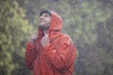 Smiling man with raincoat looking up at rain - CAIF19760